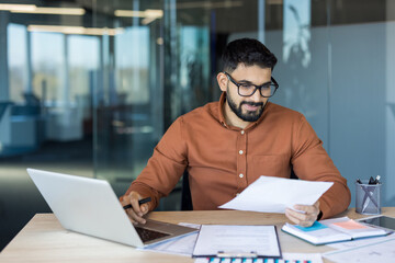 Young man in glasses and beard concentrating while working on a laptop and reviewing financial...