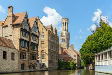 Obraz premium A row of old red brick houses and a tall Gothic bell tower in the city of Bruges, Belgium. The photo was taken from the water of a canal. The building is washed by the canal