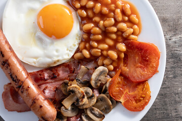 Full English breakfast with fried egg, bacon, sausages and beans on wooden table. Close up