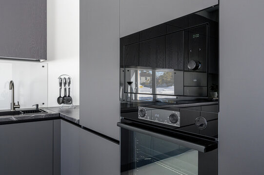 Modern grey kitchen with built-in black oven and microwave, sleek matte cabinets, dark countertop, white backsplash, stainless steel faucet, utensil holder, minimalist design.