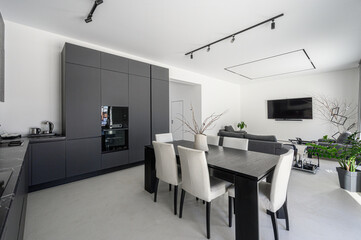 Fototapeta premium Modern open-plan space with graphite kitchen cabinetry, built-in appliances, dark dining table, white chairs, minimalist decor, track lighting, TV, greenery accents.