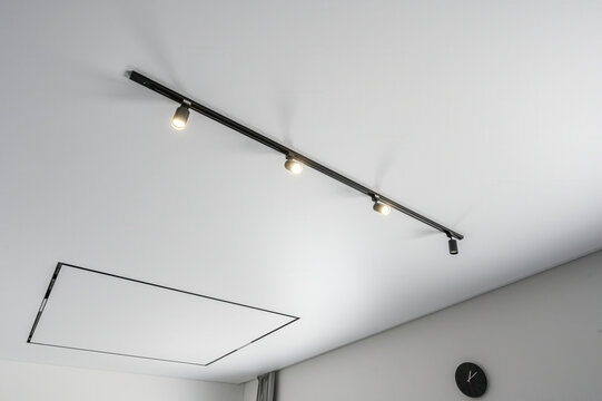 Modern ceiling with sleek black track lighting system featuring 4 adjustable spotlights; minimalist design, clean white surface, includes recessed panel and wall clock.