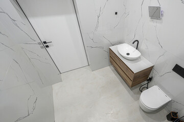 Modern minimalist bathroom with marble-patterned walls & floor, wooden vanity, oval white sink, sleek black faucet, wall-mounted toilet, clean white door, contemporary design.