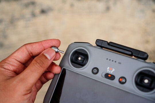 A hand holds a small metallic drone propeller near the control panel of a modern drone remote controller with visible buttons and screens in an outdoor setting.
