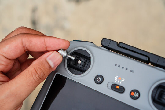 A hand carefully inserting a small metallic connector into the port of a modern drone controller with illuminated buttons and digital display showing flight mode indicators against a blurred