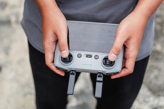 Close-up of hands gripping a drone controller with visible buttons and joysticks against a blurred outdoor background suggesting readiness for flight or operation.