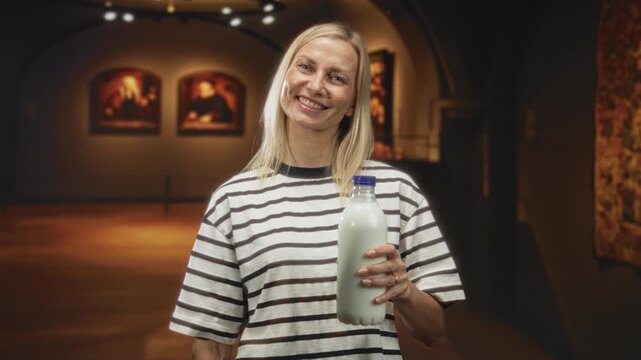 Woman holding a milk bottle with blue cap, hand visible and smiling, wearing a striped shirt in an art gallery building; serenity.