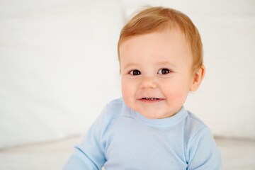 Fototapeta premium Baby close-up portrait, smiling and joyful little child on a white bed in a blue bodysuit