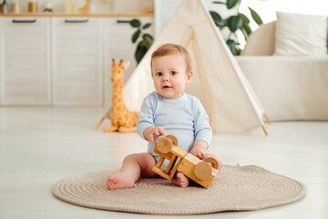 A smiling and joyful kid is playing on the floor with wooden toy cars near the wigwam in the children's room at home, a small child is sitting in a blue bodysuit laughing © Any Grant