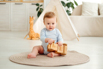 A smiling and joyful kid is playing on the floor with wooden toy cars near the wigwam in the children's room at home, a small child is sitting in a blue bodysuit laughing © Any Grant