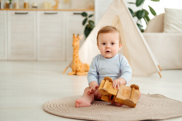 A smiling and joyful kid is playing on the floor with wooden toy cars near the wigwam in the children's room at home, a small child is sitting in a blue bodysuit laughing © Any Grant