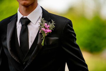 Delicate flowers of a boutonniere are captured in close-up, nestled perfectly in the black suit jacket pocket of a teenage boy or man getting ready for prom or a wedding.