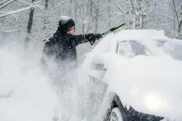 Person in black winter attire is clearing snow from a car parked in a snowy landscape, trees covered in snow visible in the background during winter weather conditions
