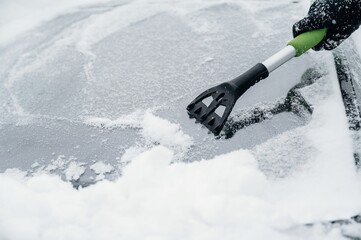 Person using a black and green snow scraper to remove snow from a car windshield covered in white...