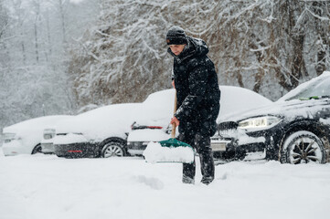 Individual dressed in black winter clothing is shoveling snow in a parking lot with parked cars covered in snow during a heavy winter snowfall scene
