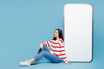 Full body side view young woman wear red striped sweatshirt casual clothes sit near big huge blank screen mobile cell phone smartphone with area isolated on plain blue background. Lifestyle concept.