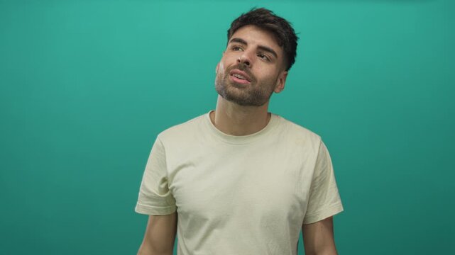 Young hispanic man raising arm to forehead gesture in white tshirt in studio against teal walls; fatigue.