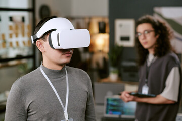 Young adult Caucasian man wearing virtual reality headset standing in modern office, while young adult man with long curly hair and glasses holding digital tablet in background observing