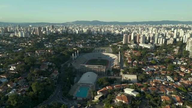 Aerial traveling shot over Pacaembu Stadium. The iconic sports arena, an outdoor swimming pool, and the dense urban horizon of S&atilde;o Paulo.