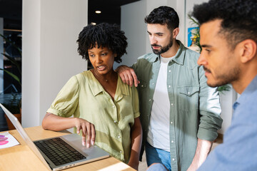 Diverse coworkers collaborating around laptop in creative office. Multicultural team analyzing...