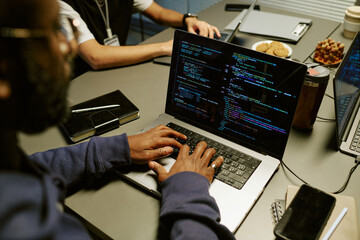 Black man typing on laptop displaying programming code, sitting at desk with notebooks and digital...