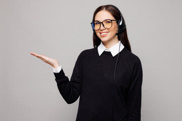 Young smiling employee operator business woman she wears set microphone headset for helpline assistance work at call center office point hand aside isolated on plain grey background studio portrait.