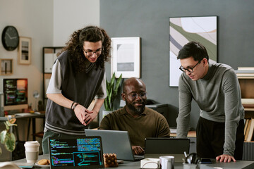 Multiethnic group of young adult men collaborating around laptops in modern office, Black man sitting and working on computer while two colleagues standing and discussing project together
