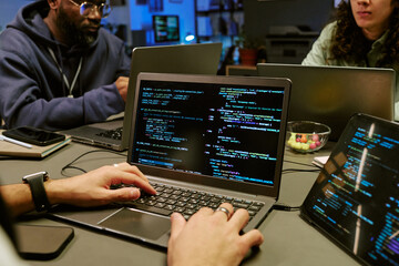 Hands of man typing on laptop displaying programming code while sitting at table with Black man and Caucasian young adult man, collaborating on software development project