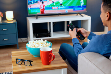 Young adult Caucasian woman sitting on sofa holding remote control and watching television with bowl of popcorn and cup on table in living room, sports game playing on TV screen