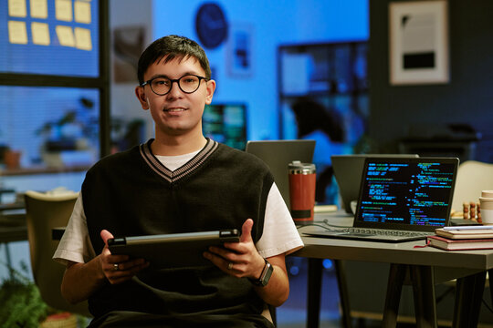 Portrait of young Caucasian man wearing glasses sitting at desk holding digital tablet, smiling at camera with open laptop displaying programming code in modern office workspace - Powered by Adobe