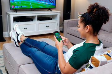 Young adult Hispanic woman reclining on sofa watching television while using smartphone, focusing on screen with legs stretched out, sports game visible on TV in living room setting