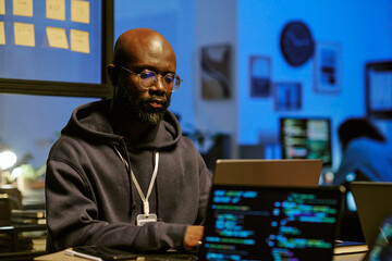 young Black man working on laptop in modern office, wearing glasses and lanyard, focusing on computer screen with programming code, sitting at desk during evening