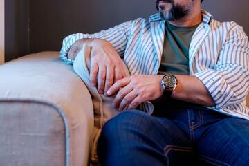 Middle aged Caucasian man sitting on sofa watching television, resting arm on cushion, wearing wristwatch, facial hair visible, relaxed posture, focused on screen