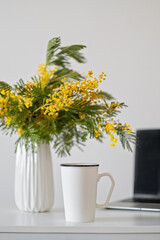Fototapeta premium White mug mockup, modern workspace with laptop and bouquet of mimosa