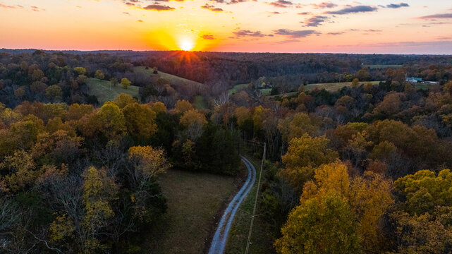 Aerial view of a winding country road cutting through autumn-kissed forests under a vibrant sunset sky, Carlisle, Kentucky, United States.