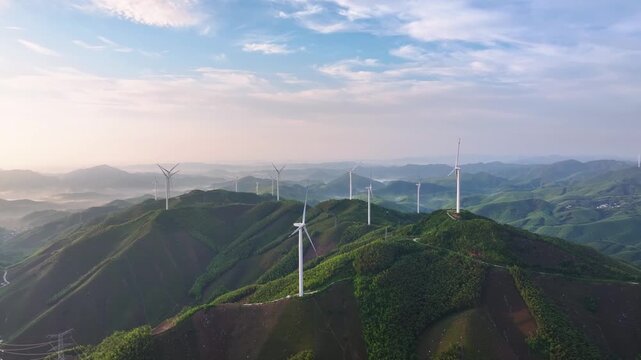 Multiple white wind turbines stand on rolling green mountains under a clear sky with morning mist