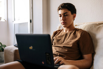 Teenage boy using a laptop while sitting on a sofa at home. Remote study, technology, digital life and modern lifestyle concept