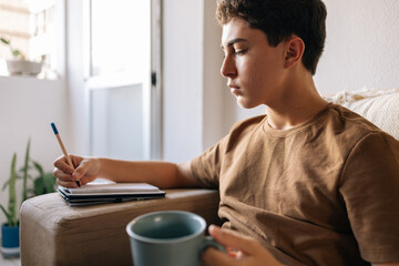 Teenage boy sitting on a sofa at home holding a cup of hot drink. Cozy lifestyle, comfort, rest, study break and quiet moment indoors