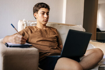 Teenage boy using a laptop while sitting on a sofa at home. Remote study, technology, digital life and modern lifestyle concept