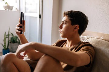 Teenage boy sitting on a sofa at home using a smartphone. Communication, social media, technology and modern teen lifestyle