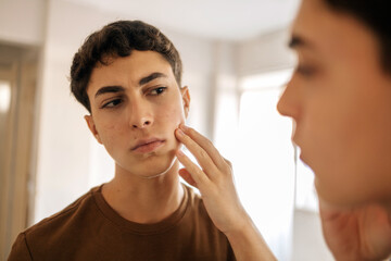 Teenage boy looking at himself in the mirror and applying skin care product on his face. Acne, self care, hygiene and adolescent routine concept