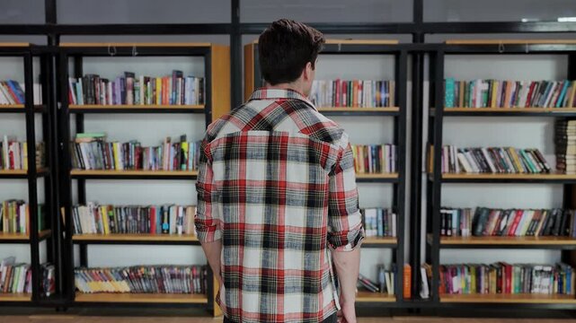 Back rear view young smart intelligent college student boy wearing casual clothes studying visit university library walk looking for novel choosing paper book standing near shelf. Knowledge concept