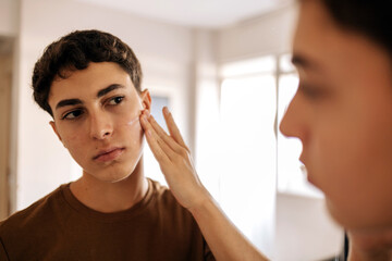 Teenage boy looking at himself in the mirror and applying skin care product on his face. Acne, self care, hygiene and adolescent routine concept