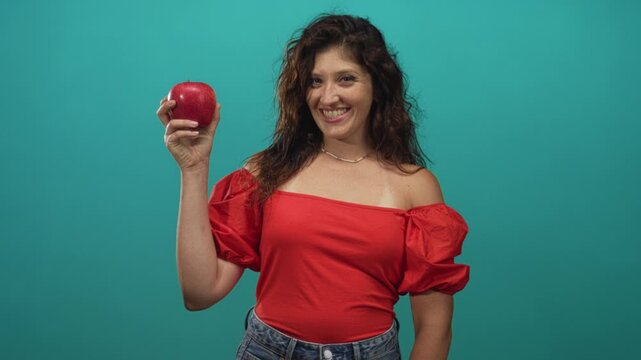 Woman wearing red top holding a red apple and smiling in teal studio with plain backdrop and half length pose; health wellness joy.