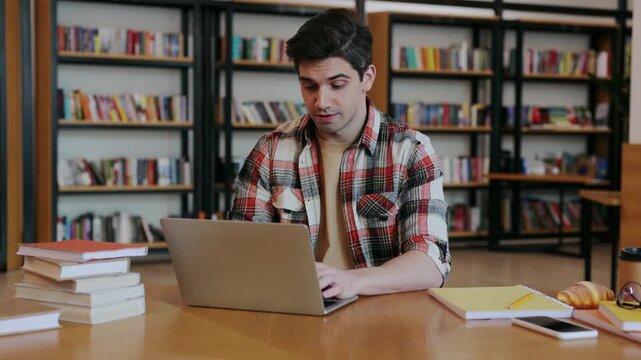 Young smart intelligent college IT student boy wearing casual clothes studying visit university library sit at desk table hold use work on laptop pc computer search information. Knowledge concept