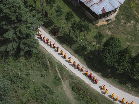 Aerial view of monks in vibrant robes processing along a winding road framed by lush greenery and traditional architecture, Gangtey Monastery, Wangdue Phodrang, Bhutan.