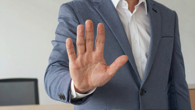 Businessman in blue suit jacket showing stop gesture with open hand to camera in office setting with computer monitor on desk in background
