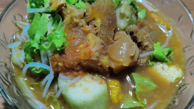 A bowl of traditional Tahu Campur Lamongan featuring beef kikil, fried tofu, rice cake, bean sprouts, and lettuce in a savory brown broth with petis sauce.