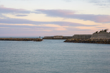 Fototapeta premium 夕焼けの海と防波堤で釣りを楽しむ人の風景。青い海と淡いピンクの空が美しいマジックアワーの景色。夏や秋の海辺のイメージ、背景素材、デザイン用。静かな漁港の夕暮れ時、趣味と癒やしのライフスタイル素材。涼しい夕方の空気感漂う爽やかな日本の海岸風景。