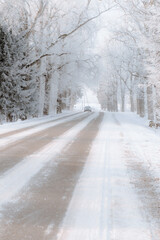 Obraz premium Car approaching on a frost-covered winter road through snowy forest, icy avenue with hoarfrost trees in soft morning light
